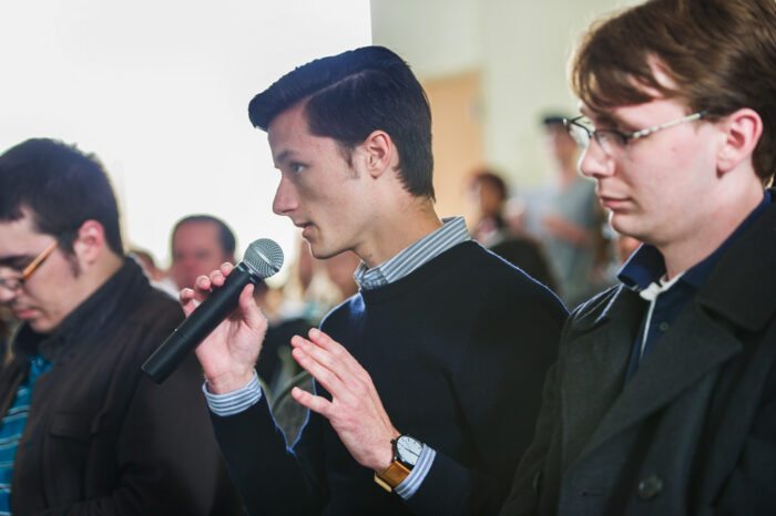 Man speaking into a handheld microphone in a town-hall style meeting.