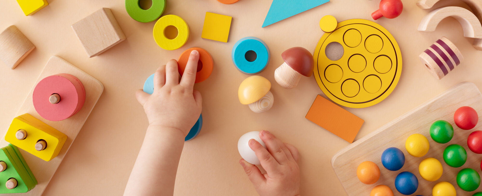 A child's hands are playing with colorful wooden educational toys, including geometric shapes, stacking blocks, and puzzle pieces on a beige surface.