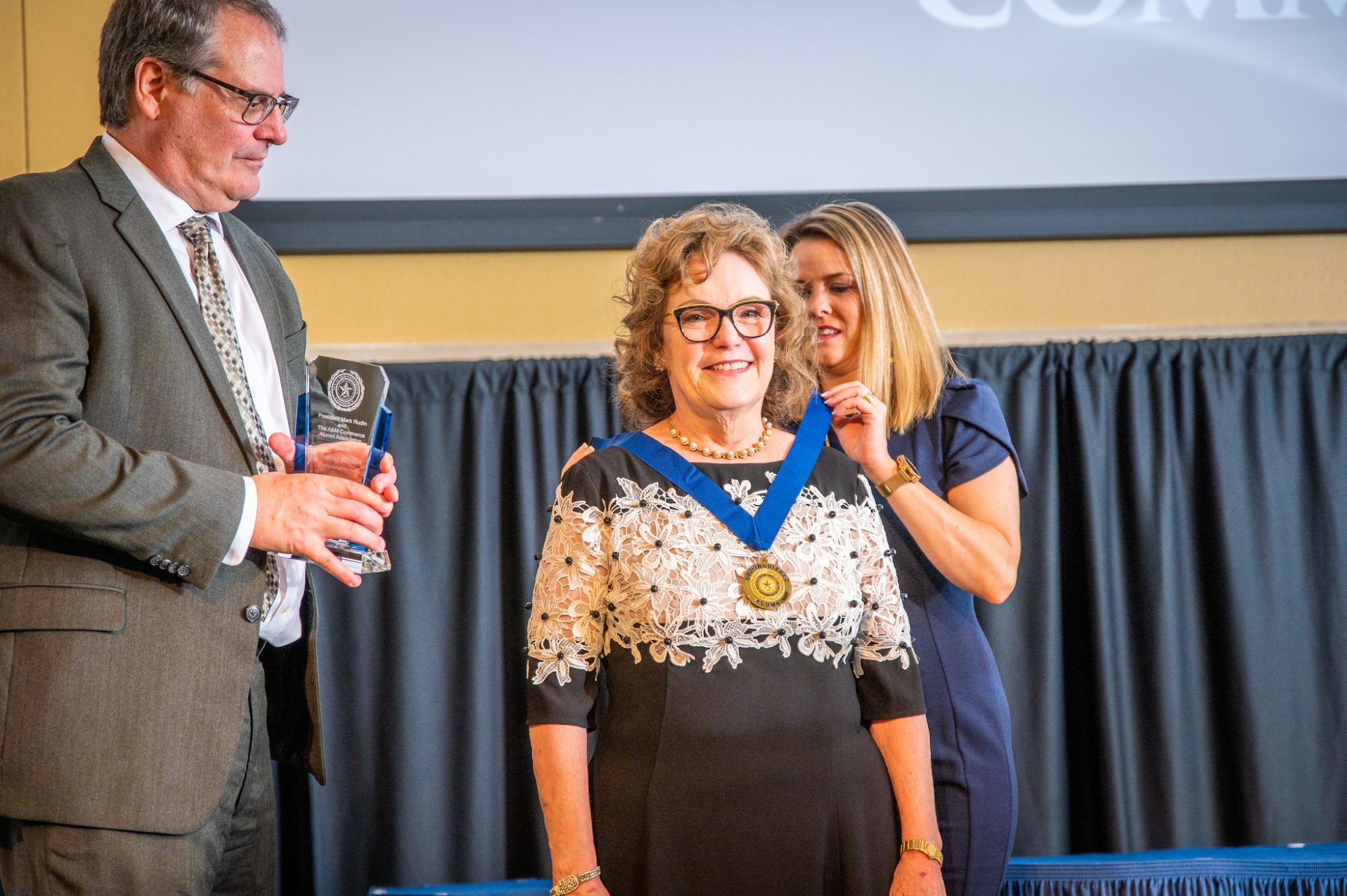 Awardee allows a medallion to be fastened around her neck, with presenters on either side of her.