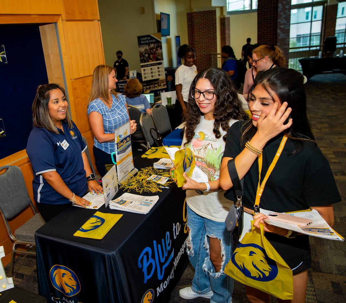 Students smile for the camera in front of a table holding brochures. The table has a black cloth with "Blue to Gold Money Management Center" written on the front.