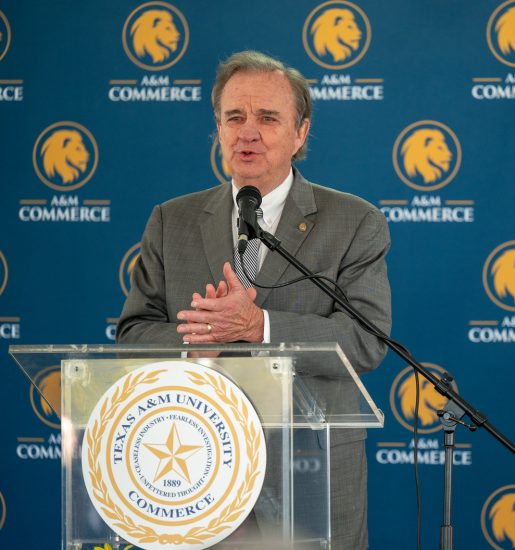 Chancellor John Sharp speaks behind a podium with hands clasped. A blue backdrop with the A&M-Commerce logo is behind him.