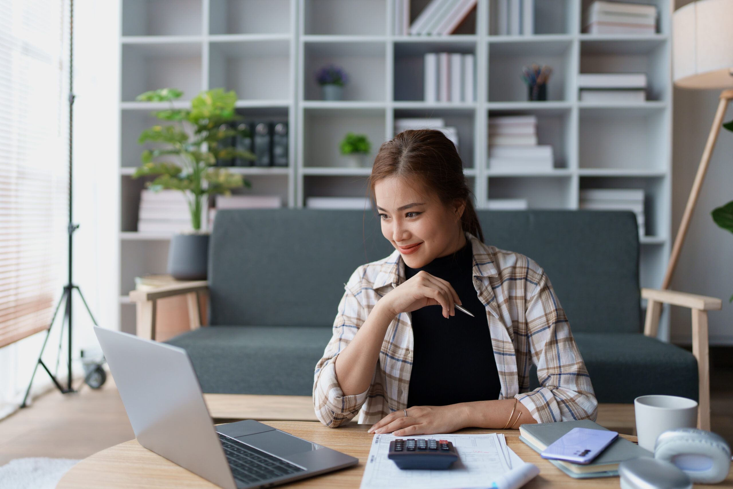 A woman sits at a table working on her laptop in a modern living room. She is smiling while holding a pen, with a calculator and documents in front of her.