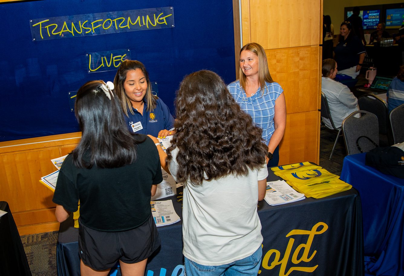 Two money coaches stand behind a table, assisting two students in front of the table, which has flyers on top of it.