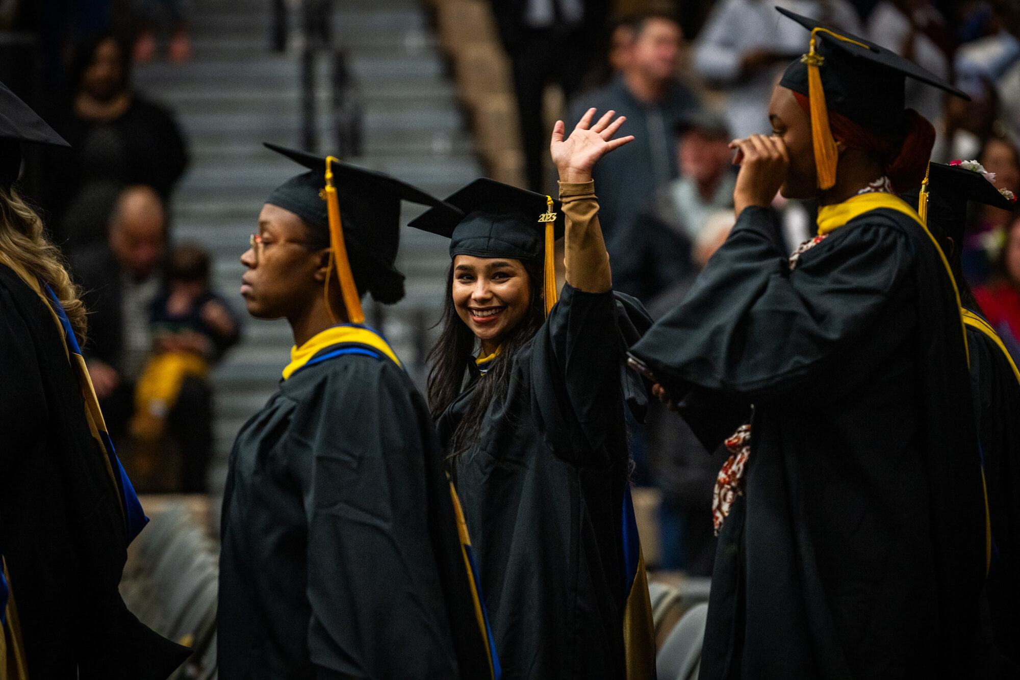 Student at graduation ceremony waves at the camera, wearing black gown and black graduation cap. Graduates walk in front of and behind her.