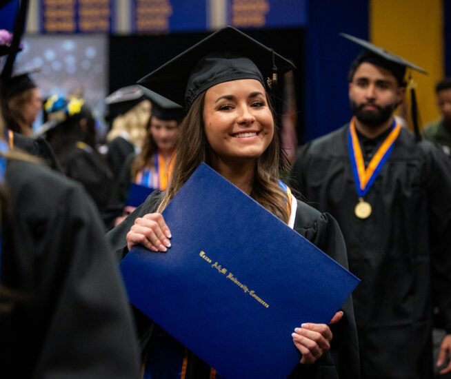 Graduate holds diploma in both hands and smiles at the camera, wearing graduation cap and gown.