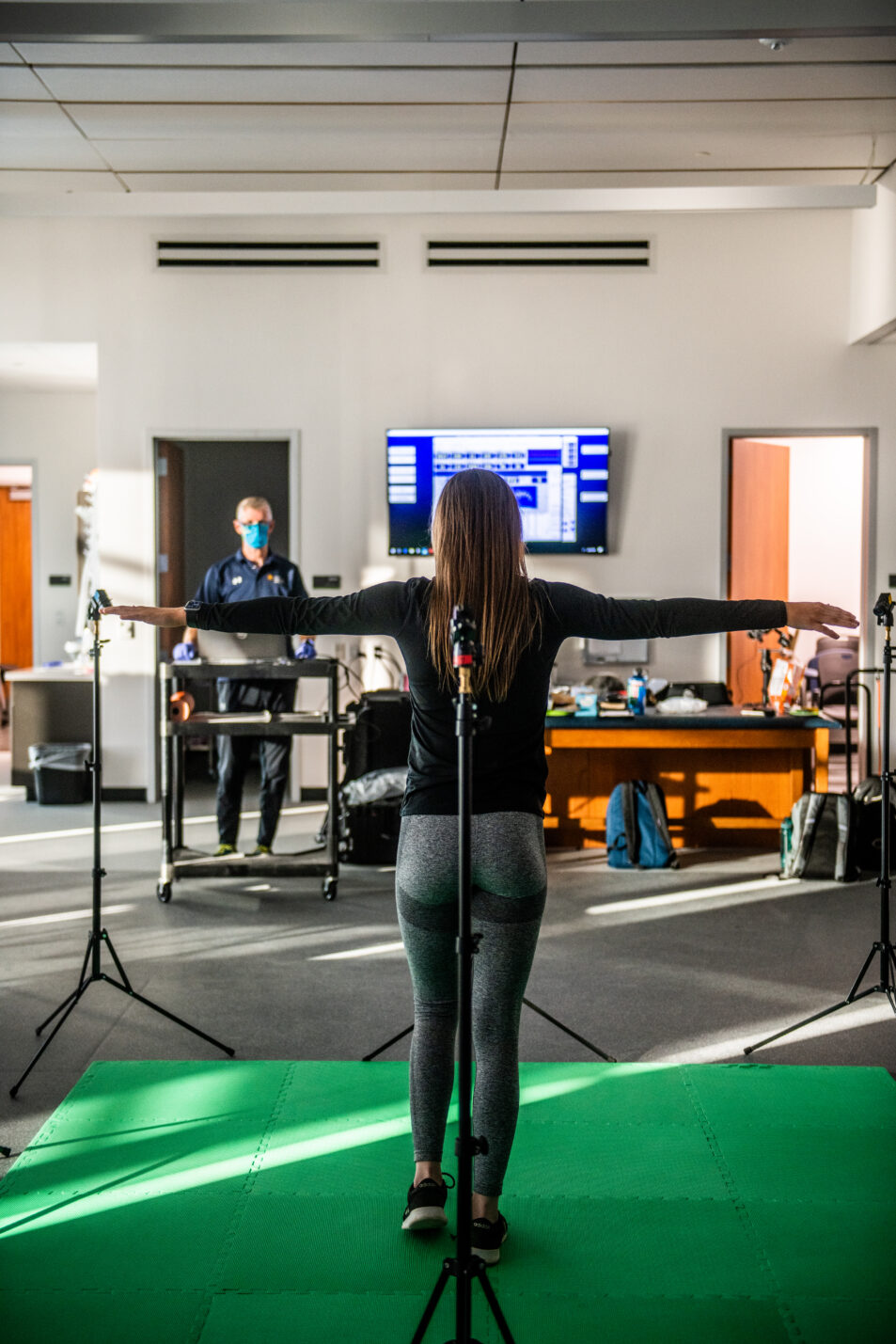 A woman stands facing away with arms raised as part of a physical assessment conducted by a faculty researcher.