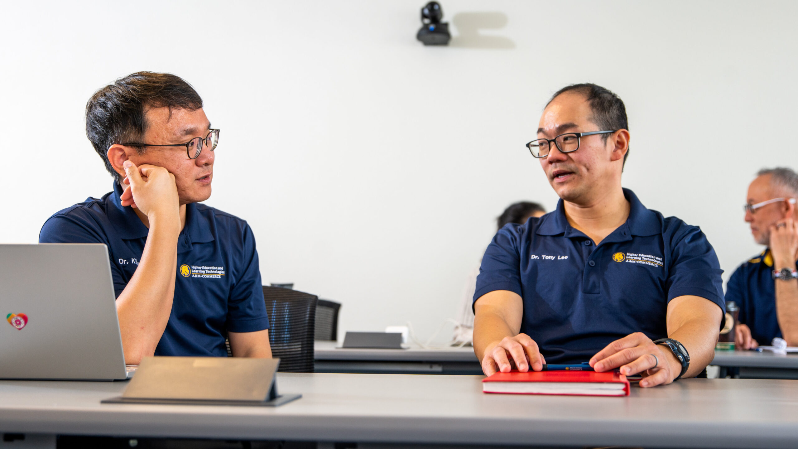 Two men in navy blue polo shirts sit at a table in a meeting. One listens with his hand on his face, while the other, holding a red notebook, speaks. A laptop is in front of them.