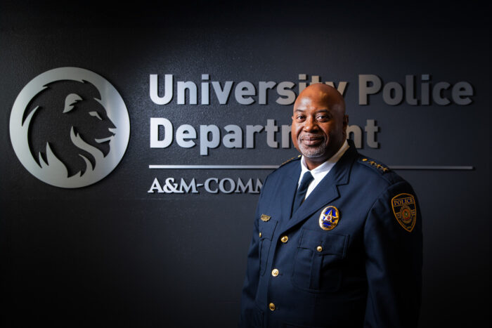 Chief Bryan Vaughn dressed in his dark blue, formal police uniform. He is standing in front of a dark wall with a silver embossed TAMUC lion head logo and the words University Police Department A&M-Commerce.