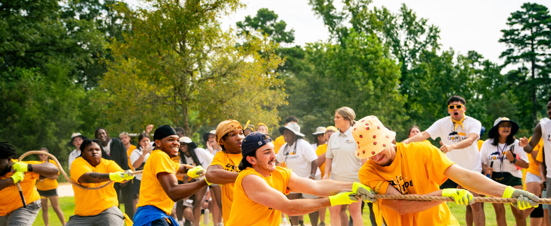 Students play tug-of-war on sand, wearing gold shirts. The audience cheers in the background.