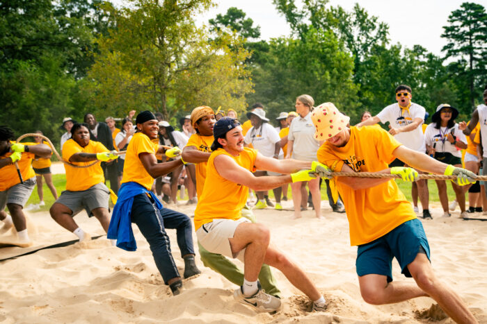 Students play tug-of-war on sand, wearing gold shirts. The audience cheers in the background.