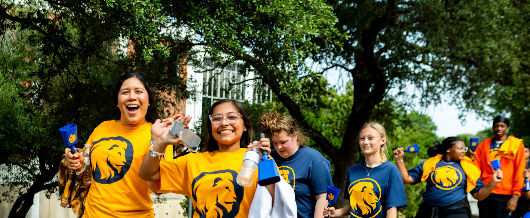 Students walk down the sidewalk on campus, smiling at the camera. Two students in front wear gold t-shirts with the school's lion logo on the front. The students are flashing the "L" hand sign as a show of school spirit.