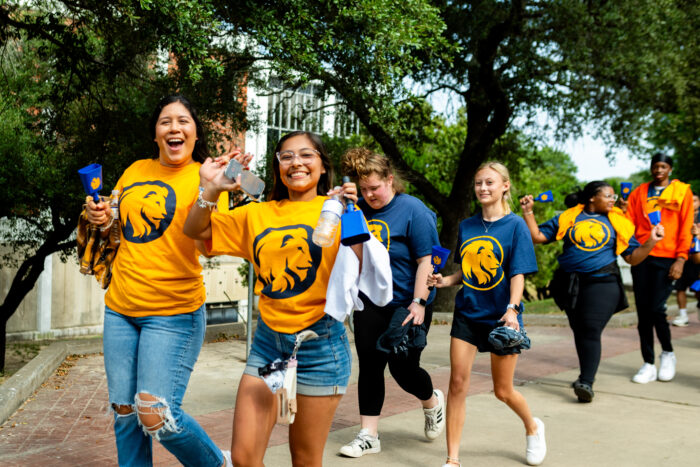 Students walk down the sidewalk on campus, smiling at the camera. Two students in front wear gold t-shirts with the school's lion logo on the front. The students are flashing the "L" hand sign as a show of school spirit.