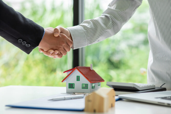 Two people shake hands, presumably over a real estate deal, with a table that contains a small model of a home beneath their hands.