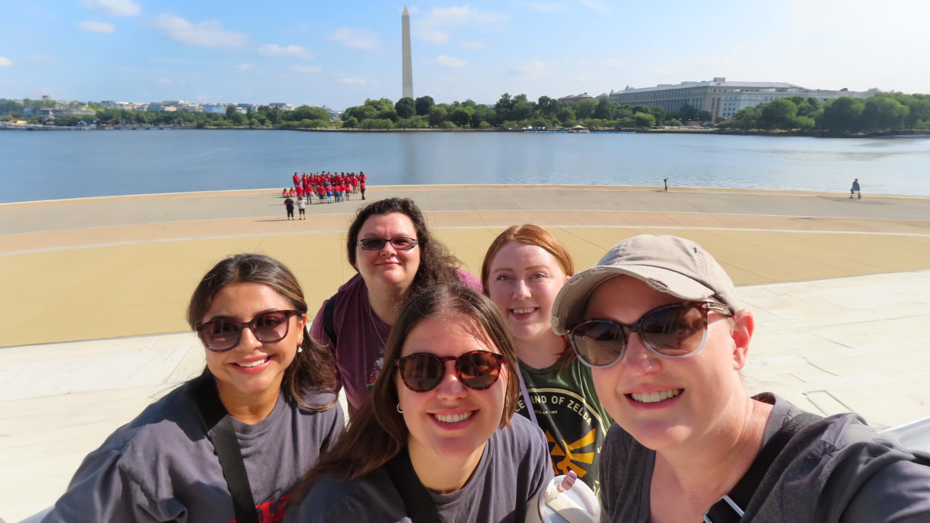 A woman wearing a cap takes a selfie with four female college students. Behind them, a group of sightseers in red shirts stand at the edge of the Tidal Basin with the Washington Monument standing tall in the distance.