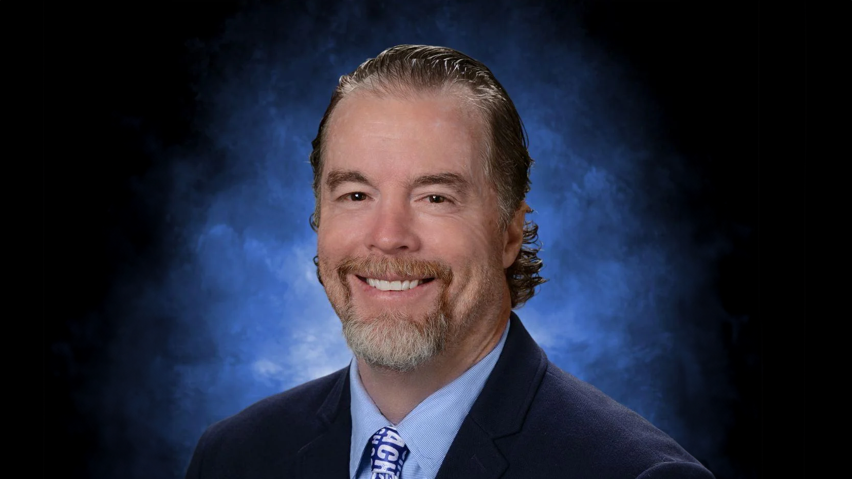 A professional headshot of A&M-Commerce alum Shae Creel in a dark suit and blue tie against a blue and black backdrop.