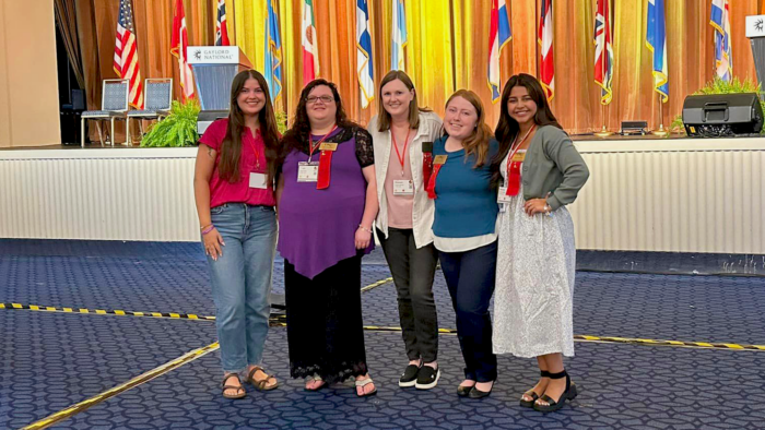 Four smiling female college students pose with a female faculty member in the middle. A stage behind them is adorned with flags representing countries around the world.