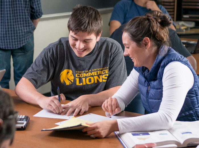 Two students work together at a table in a classroom. A student in the background is at the blackboard.