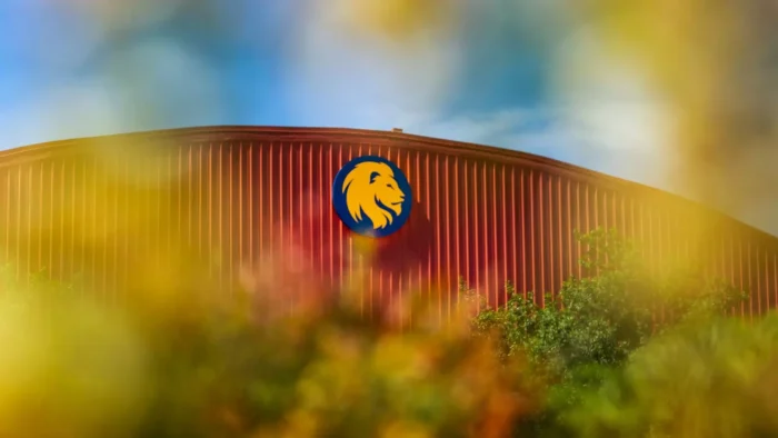 Dome-topped fieldhouse peaks out from behind trees. Lion head logo is placed at the top of the building.