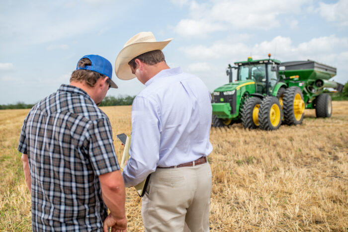 Two men, one in a cowboy hat and another in a blue cap, are standing in a field beside a large green tractor. They appear to be discussing something while looking at a cell phone.