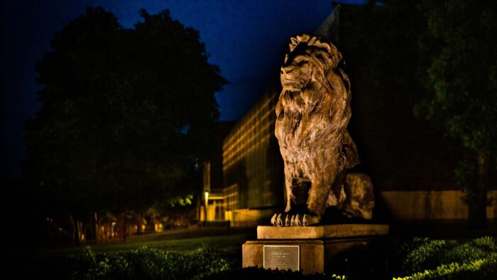 Lion statue is in a sitting pose. Photo is taken at night with statue lit up.