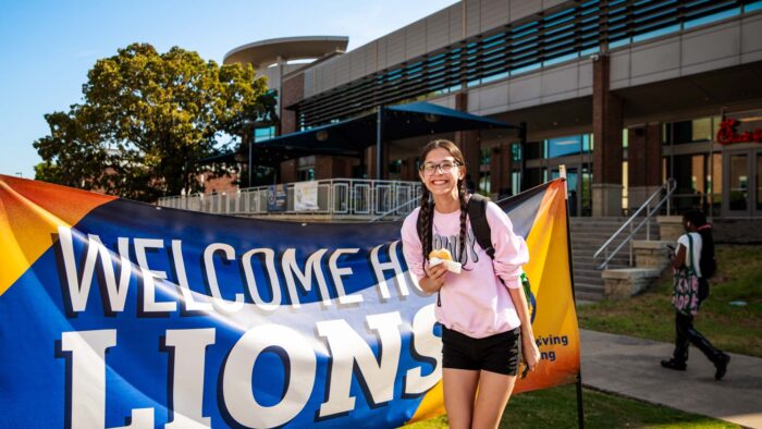 Student poses in front of a "Welcome Home Lions" banner, wearing shorts and a t-shirt and holding a donut.