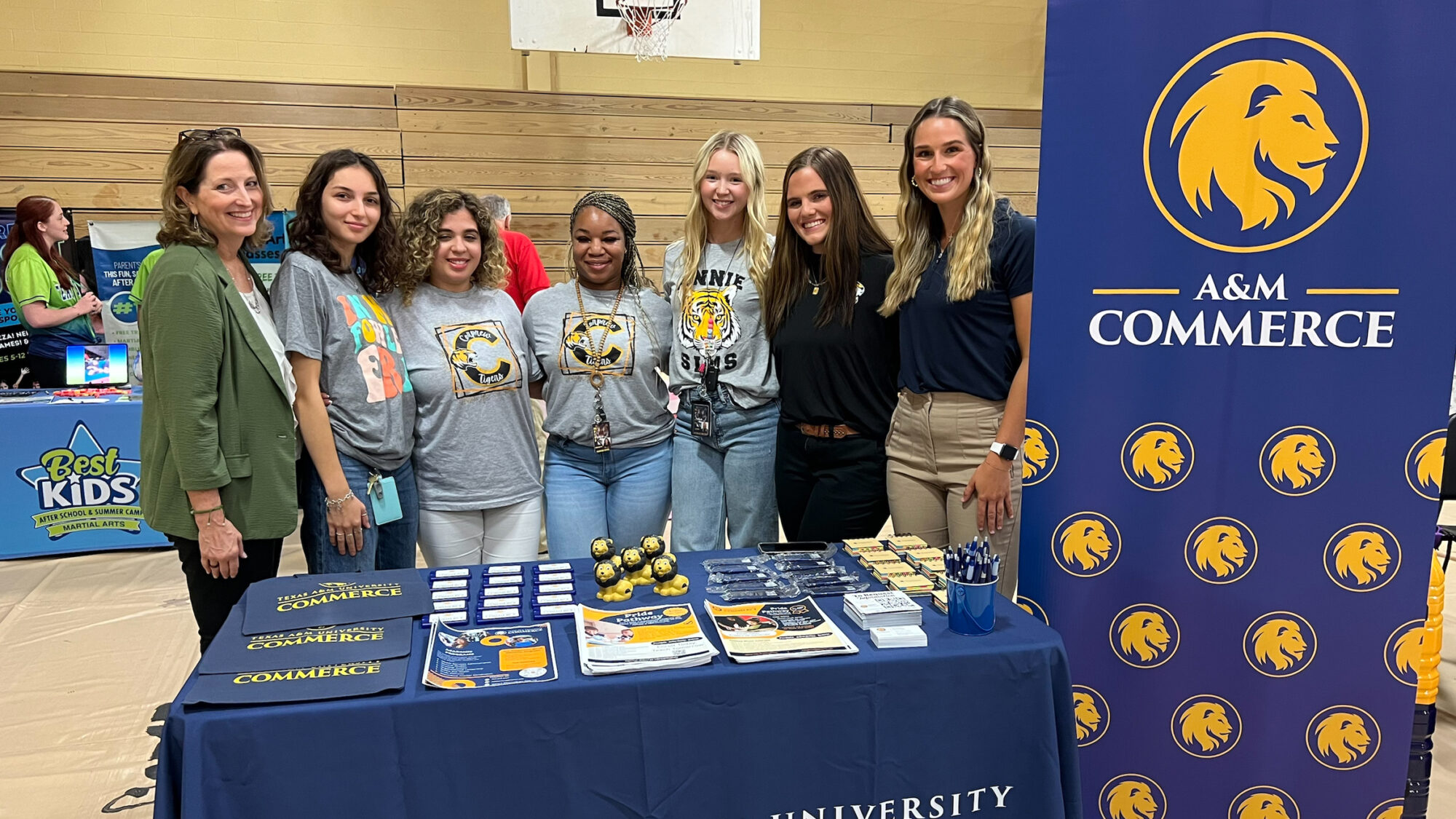 Five teacher certification scholarship recipients flanked by two TAMUC representatives stand shoulder-to-shoulder in front of a table with a blue cloth cover and promotional items on top. Next to them is a blue, vertical banner with the A&M-Commerce logo and numerous smaller lion head logos below.