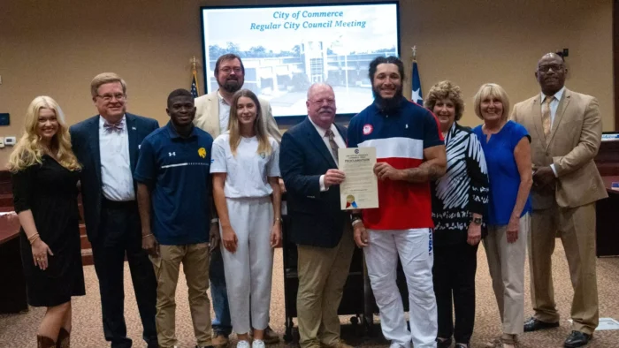 Ten individuals face the camera and smile, with a screen in the background that reads "City of Commerce Regular City Council Meeting."