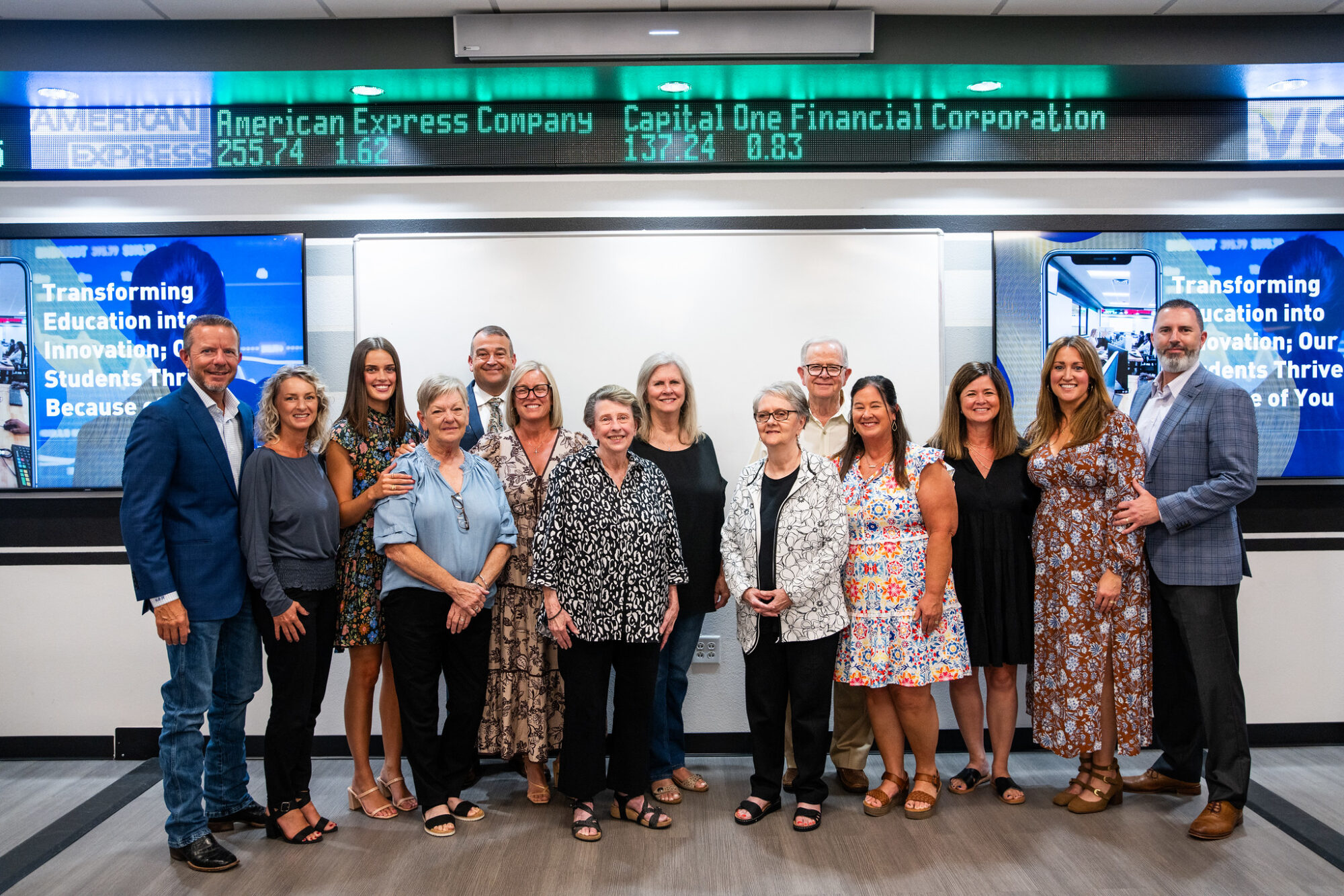 Fourteen family members face the camera and smile. Behind them there is a white board flanked by two computer screens with blue backgrounds and quote written in white. The quote reads "Transforming education in innovation; our students thrive because of you.