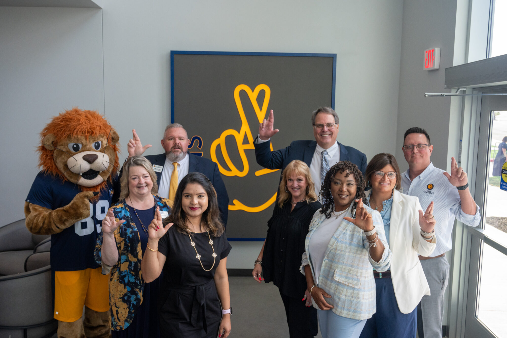 Eight A&M-Commerce employees and Lucky the lion mascot face the camera and hold up their hands in an "L" shape in a show of school price. They stand in front of a graphic representing the "L" hand sign.