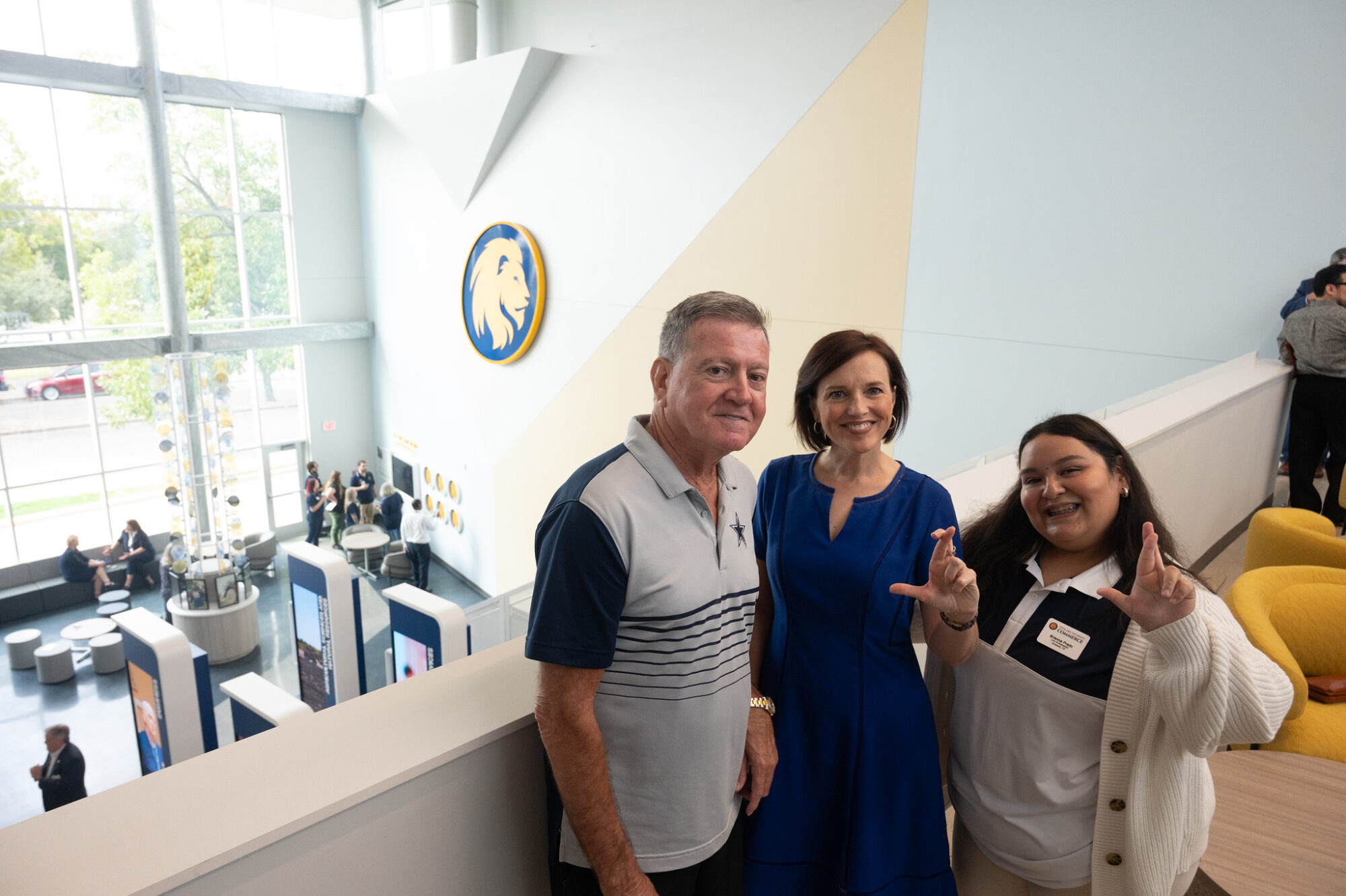 Three ribbon-cutting attendees stand on the second-floor balcony. Two of them hold their hands up in an "L" shape as a sign of school pride. All of them are smiling.