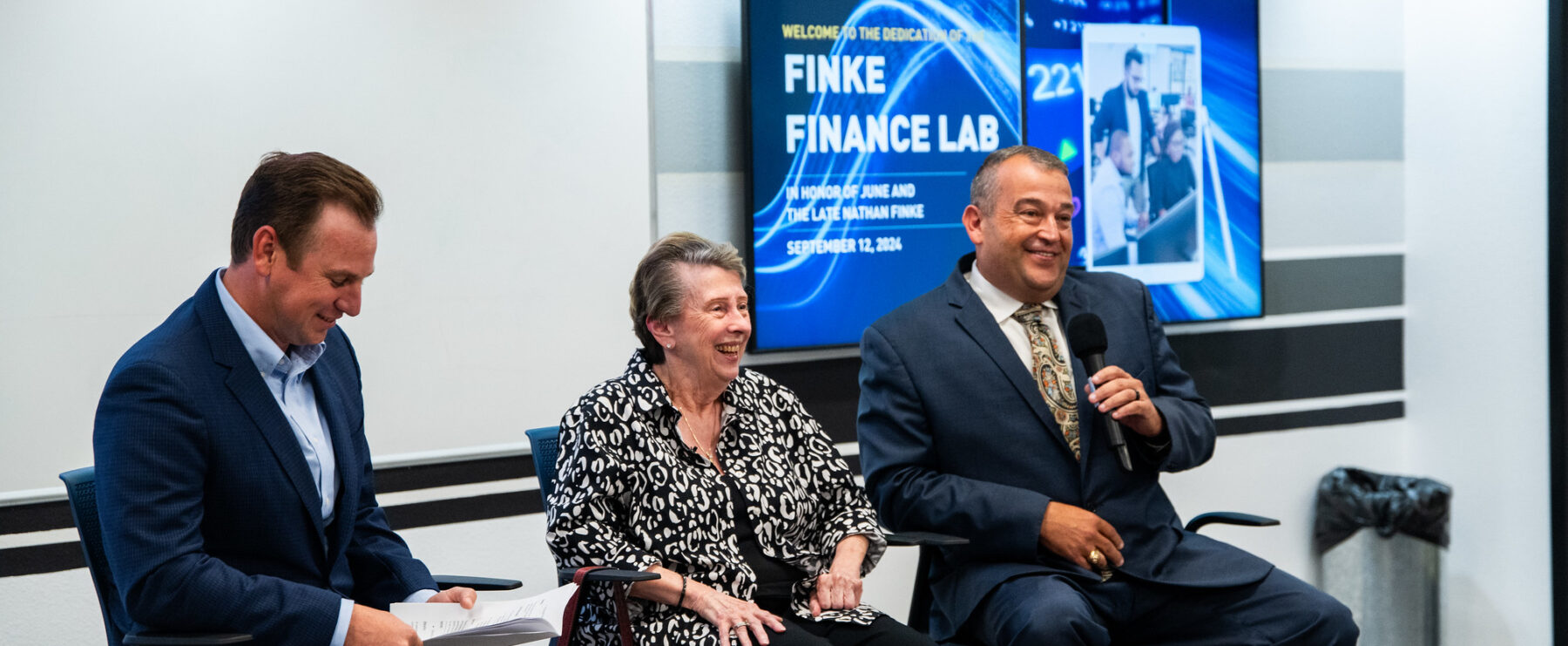 Three panelists sit at the front of the room in raised chairs. The panelists smile at the audience.