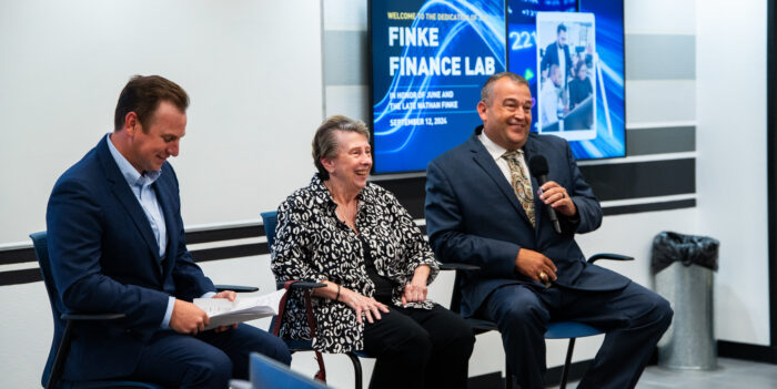 Three panelists sit at the front of the room in raised chairs. The panelists smile at the audience.