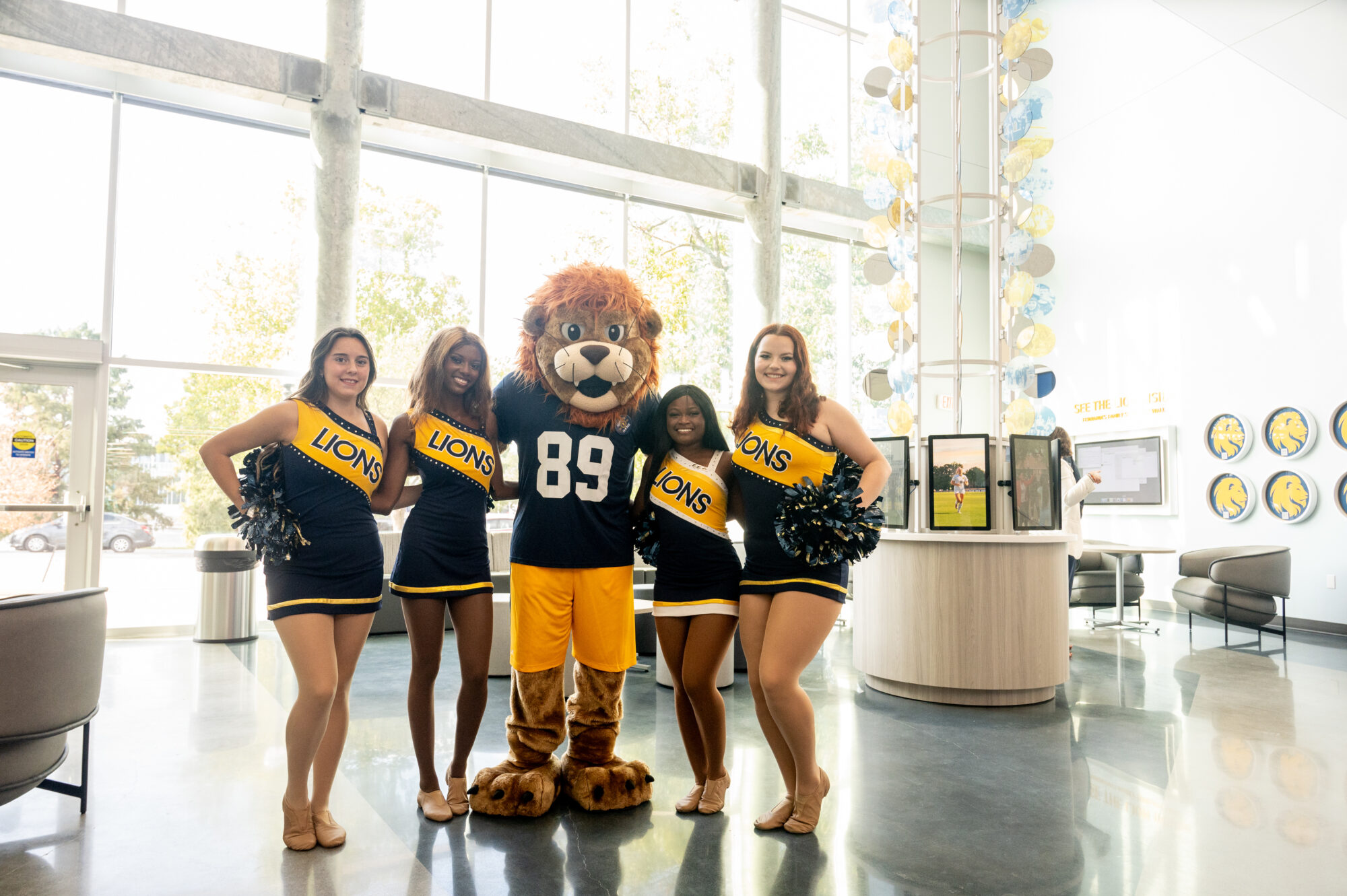 Lion mascot stands in Welcome Center with two cheerleaders on each side of him.