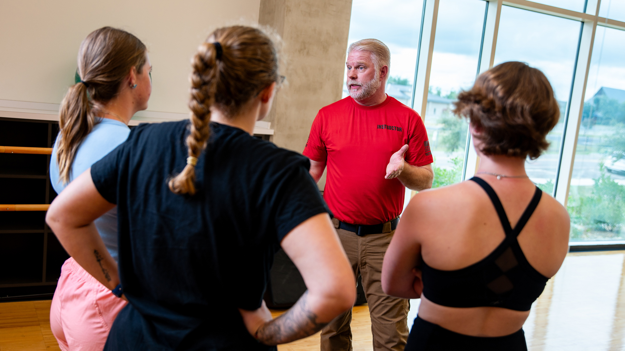 An instructor discusses defense tactics to three students who are facing away from the viewer.