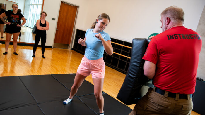 A student with raised fists prepares to demonstrate a punching technique. An instructor stands holding a thick safety pad to absorb the impact. Two students watch a few feet away.