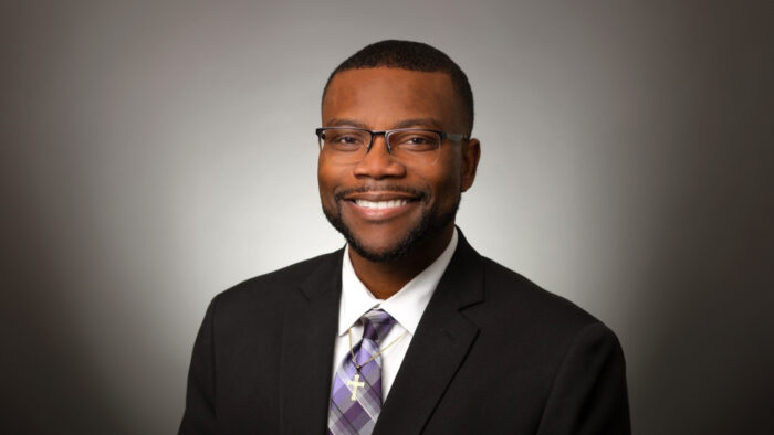 Dr. Winston McCowan poses for a headshot against a vignette background wearing a dark suit, purple-toned tie and a gold cross necklace.