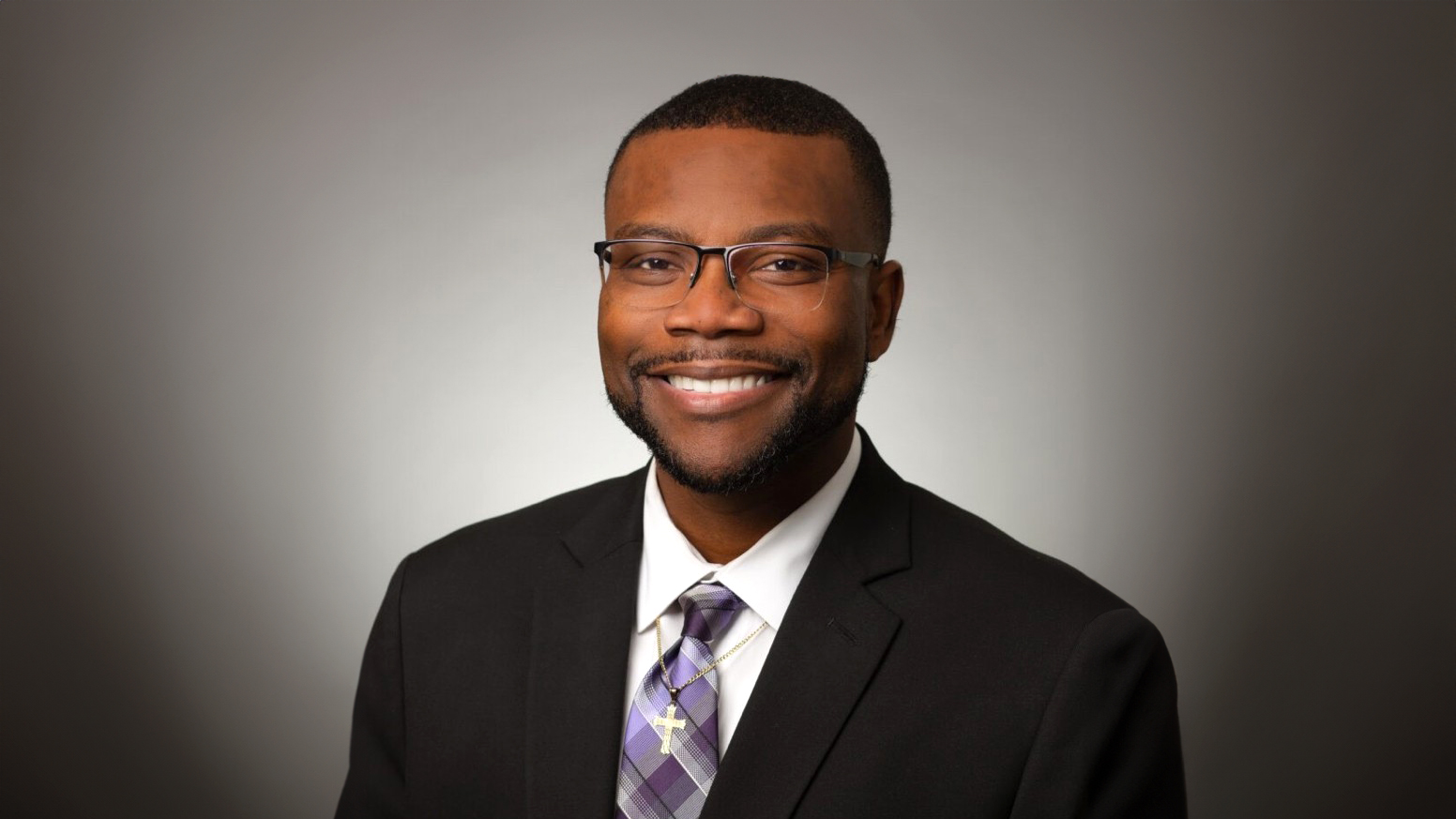 2024 ESC Region 11 Principal of the Year Dr. Winston McCowan poses for a headshot against a vignette background wearing a dark suit, purple-toned tie and a gold cross necklace.