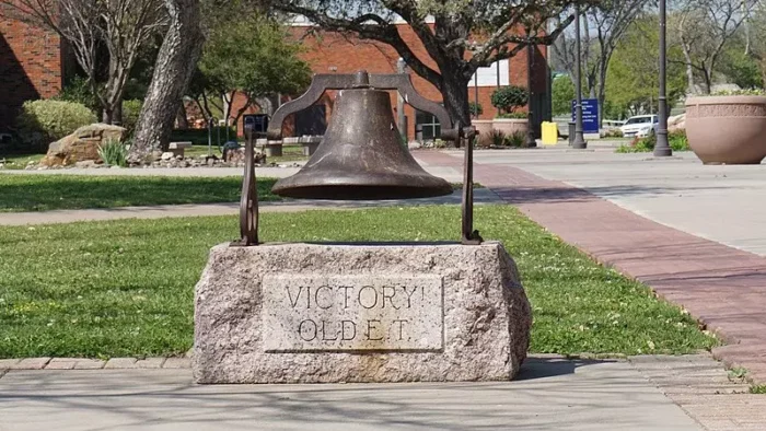 Large metal bell is mounted on a stone base. University campus building is shown behind the bell.