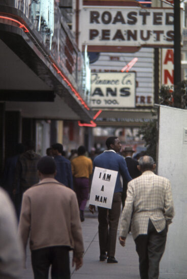 A photo from 1968 showing people walking down a downtown street. One person is wearing a sign on their back that reads "I am a man."