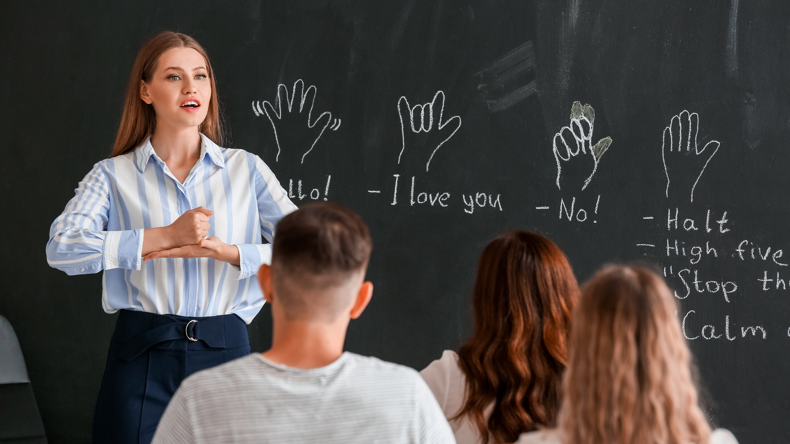 A bilingual teacher stands at the front of a classroom demonstrating ASL. Behind her is a blackboard with chalk drawings of ASL signs.