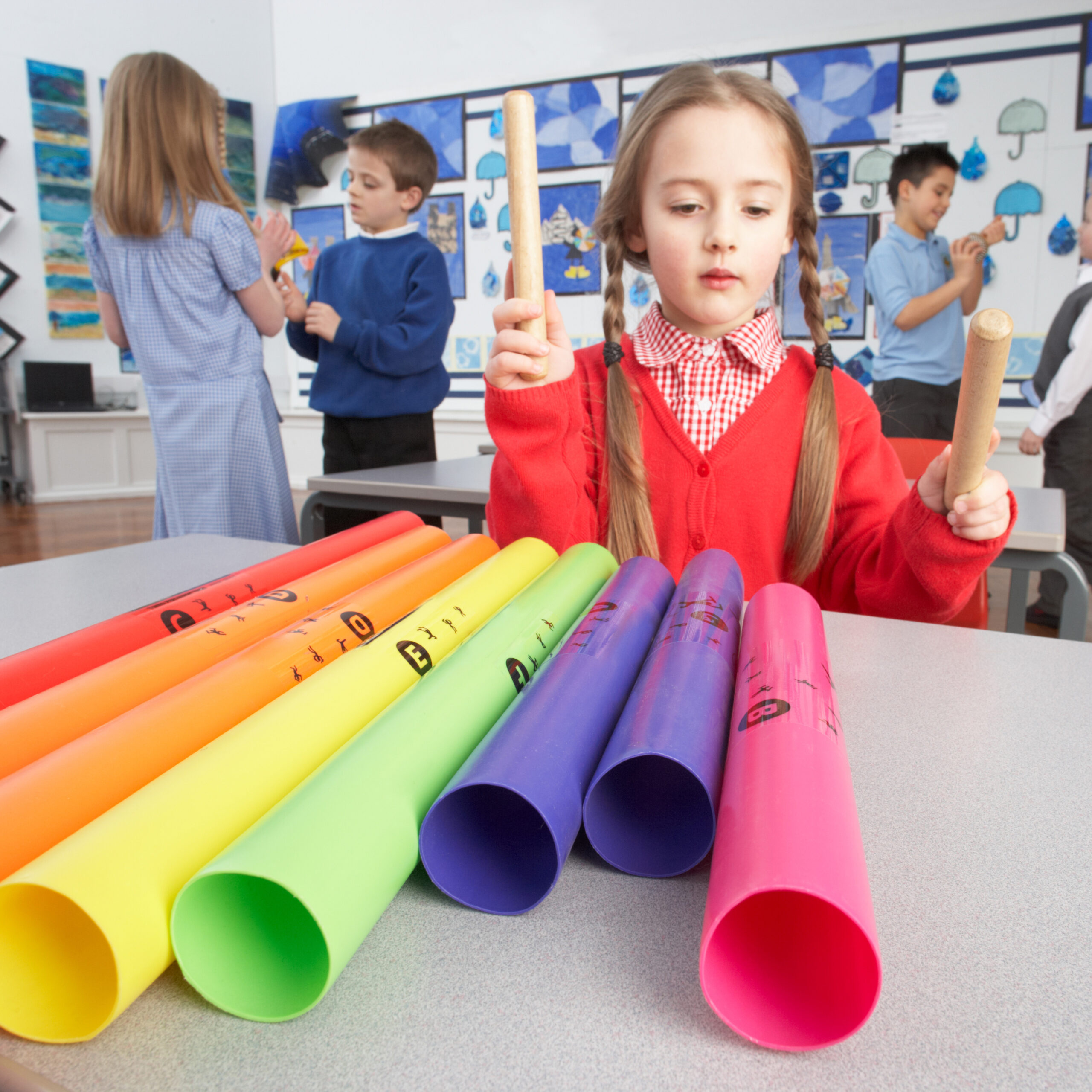 A young girl in a red sweater plays Boomwhackers in a classroom, tapping rhythm sticks on colorful plastic tubes in front of her.