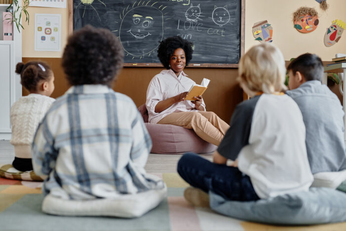 Teacher sits at the front of a classroom reading a book to several students who are sitting on cushions.