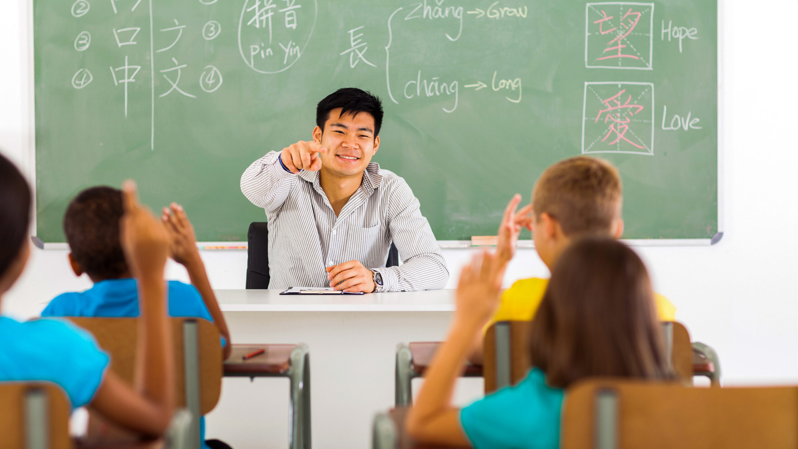 An instructor sitting at a desk at the front of a classroom points toward students with hands raised in the foreground.