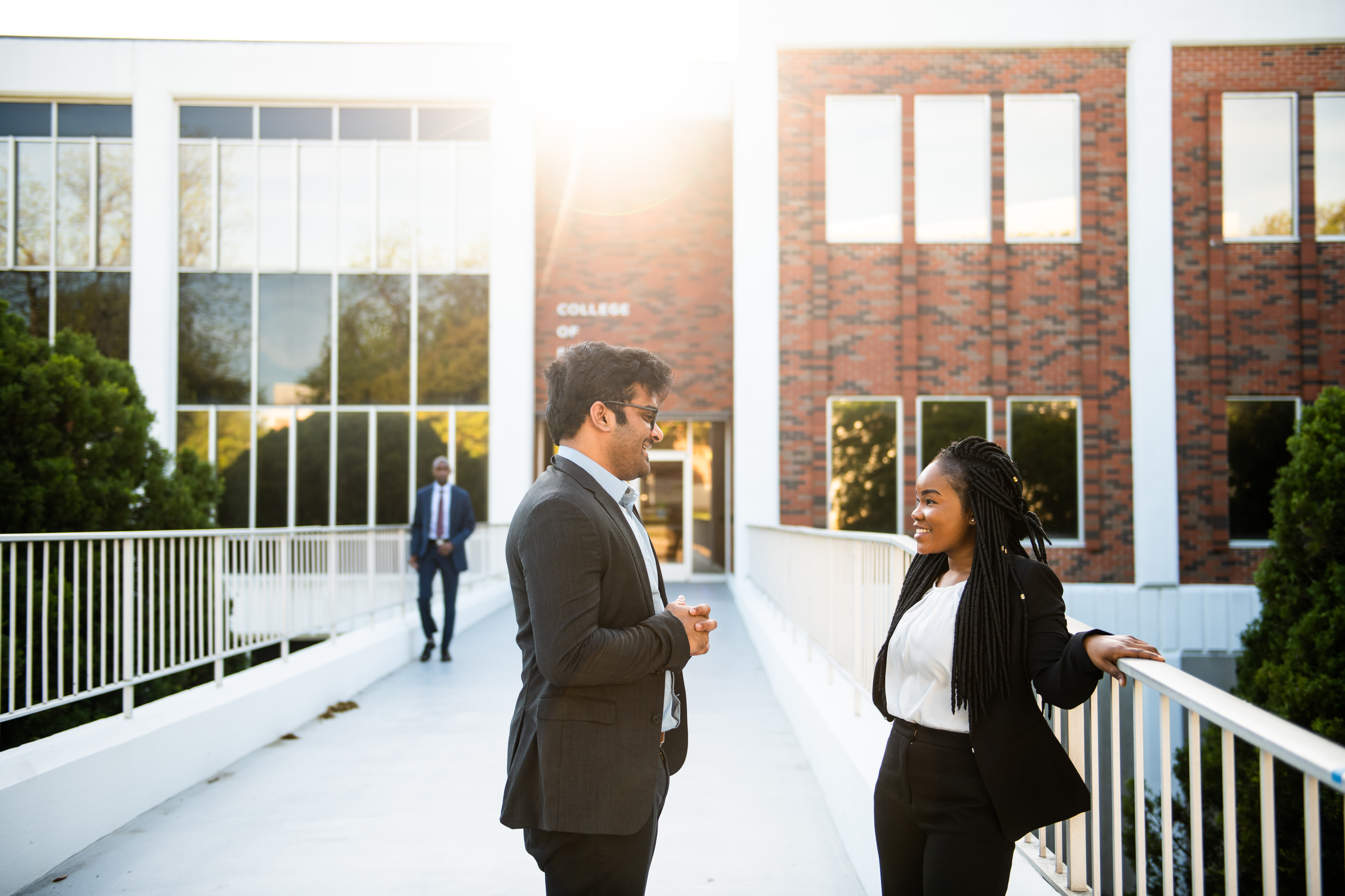 A man and a woman, both wearing business attire, engaged in conversation while standing on a walkway in front of a brick building in the evening light.