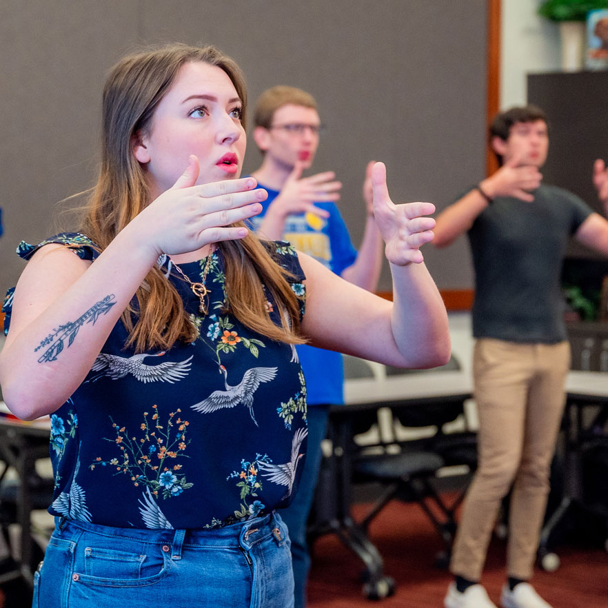 A young woman in a floral shirt gestures with her hands, focusing intently during a group activity, as others follow her movements.