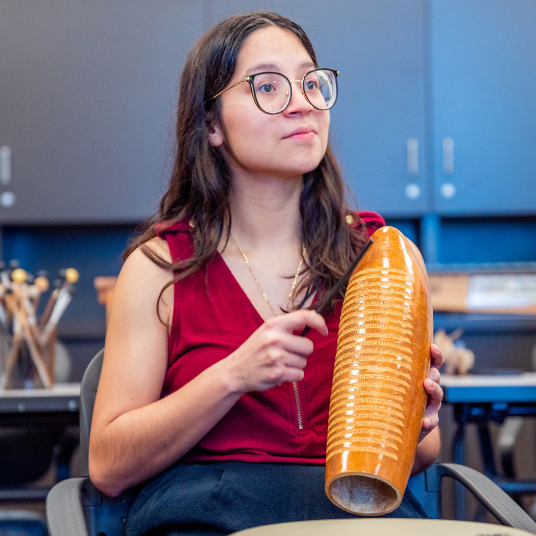 A young female wearing glasses and a red sleeveless shirt is looking forward and holding an instrument. The background appears to be in a classroom.