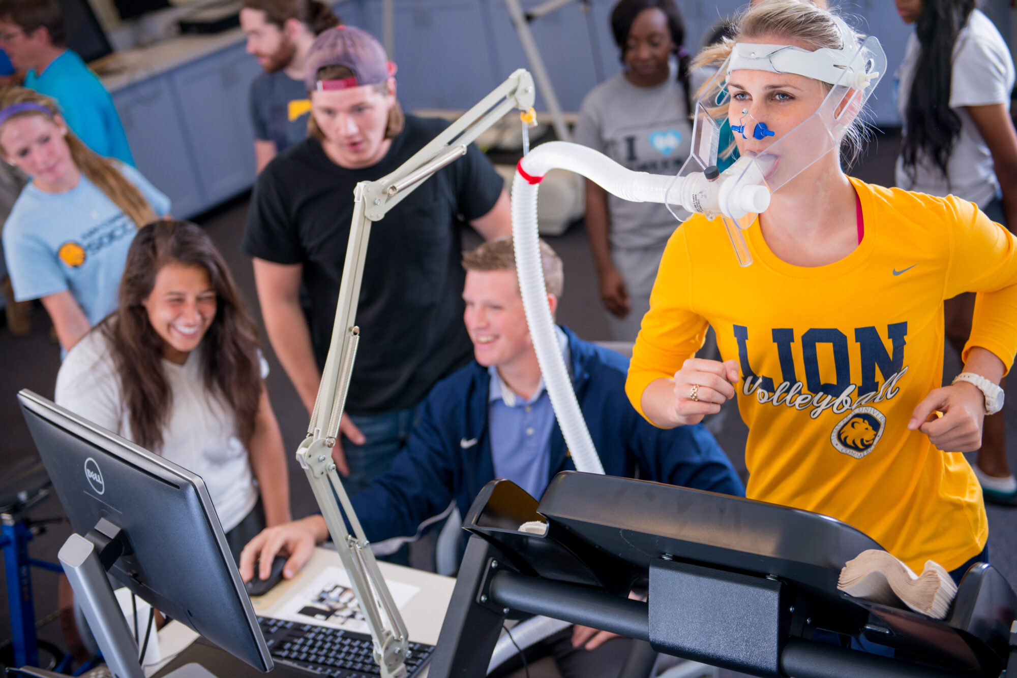 An athlete runs on a treadmill while connected to a tube measuring her breathing. Several students in the background are analyzing the data on a computer monitor.