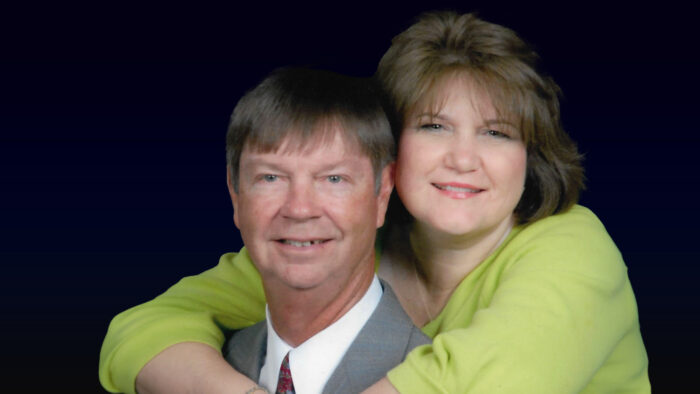 A head-and-shoulders portrait against a dark background of a man in a gray suit with a woman in a green top embracing him from behind with her arms and hands crossed over his shoulders.