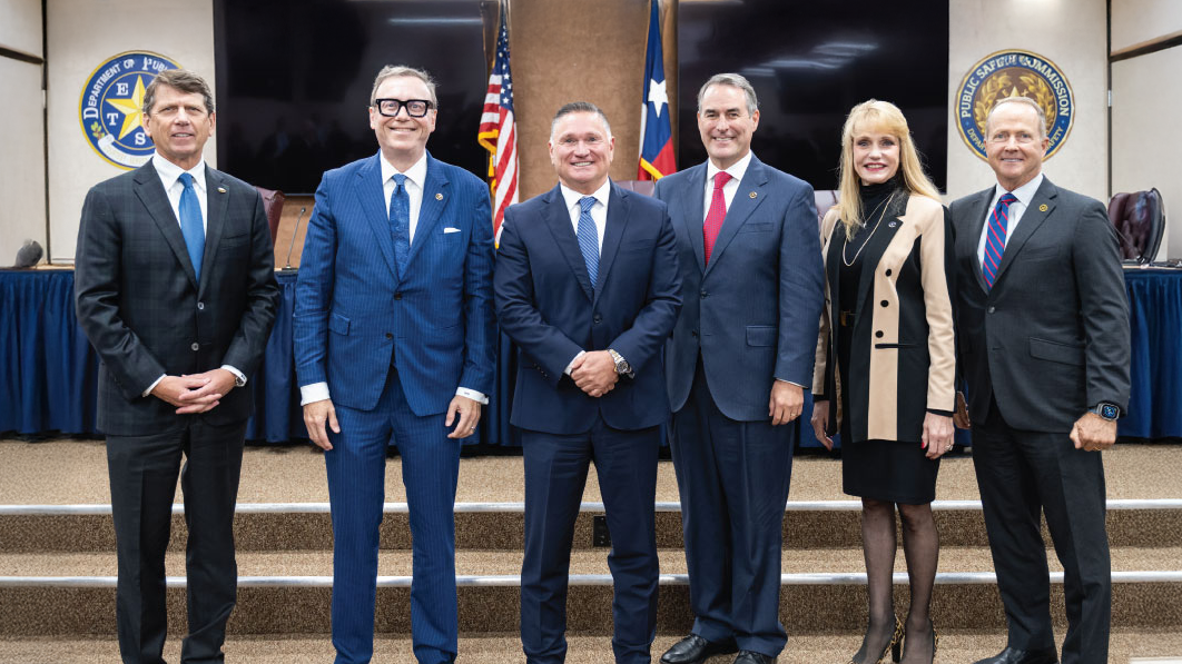 ETAMU alum Freeman Martin is flanked by two men on his right and a woman and a man on his left. All are wearing formal suits standing side by side for a photo. Behind them are steps leading to a short stage with the American and Texas flags in the background.