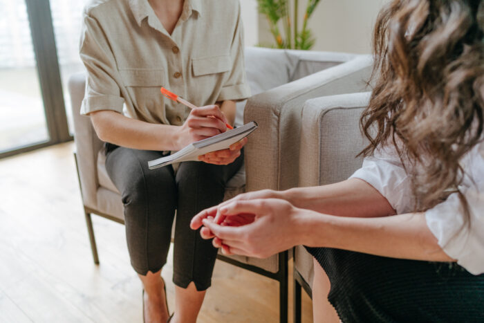 Two people sit sin chairs, representing a counseling session. Their faces are not in the frame. The photo shows the counselor writing on her notepad in the background and the patient with her hands clasped together on her lap in the foreground.